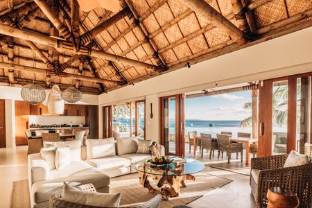 A wide-angle view of the inside one of Kokomo’s luxury residences, showcasing its traditional Fijian design-inspired high thatched ceiling, large living room space and adjacent modern kitchen, with the ocean visible directly outside the wide open doorways and across its expansive deck.