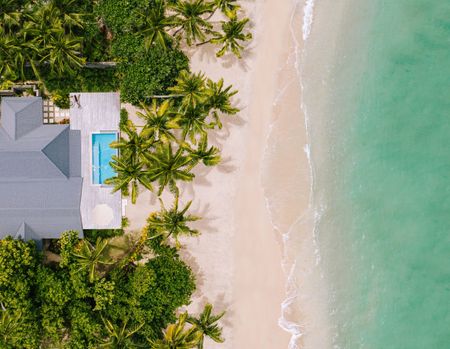 A birds-eye view of a Kokomo beachfront villa and the adjoining pristine white sandy beach and clear turquiose water. The villa’s roof and private deck with an infinity pool are visible between numerous palm trees and other vegetation.