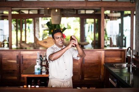A smiling Kokomo bartender standing behind one of Kokomo’s bars while shaking a cocktail shaker, with a large hibiscus flower tucked behind their ear. A wide public area is visible behind the bar.
