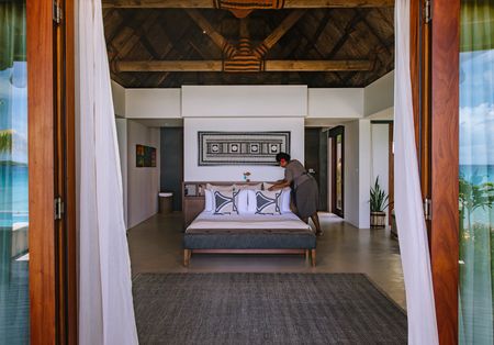 Looking in from the deck into the bedroom of of Kokomo’s villas while a member of Kokomo’s housekeeping team is making up the bed. A large piece of Fijian artwork is hanging above the bed.