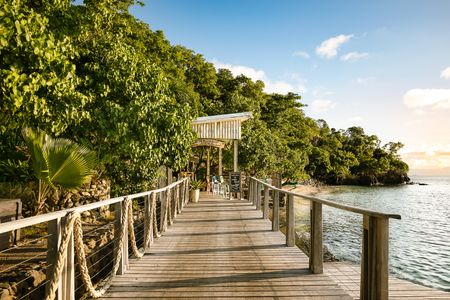 A wooden walkway alongside the ocean leading to the entrance of one of Kokomo’s ocean-front restaurants nested in amongst lush vegetation as the sun is beginning to set.