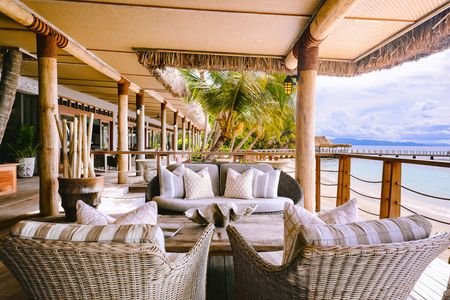A large sofa and two large wicker armchairs around a rustic timber coffee table with a large shell-shaped centrepiece on a shaded deck extending over the sandy beach, adjoining one of Kokomo’s beachfront restaurants.