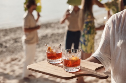 Person standing on the beach holding a serving board with two cocktails sitting on top.