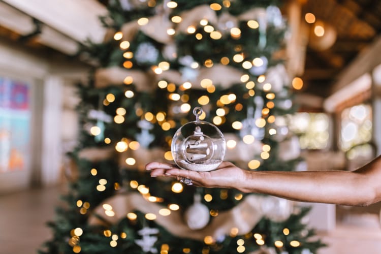 A person holding a glass bauble containing small hard-carved wood pieces atop a bed of beach sand, being held in front of a lit up Christmas tree in the Kokomo Island Fiji welcome centre.