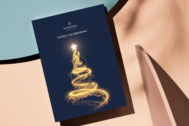 A dark-blue printed booklet with Kokomo Private Island Fiji Festive Season 2023 on the front and a large gold Christmas tree graphic made up of various snowflake shapes, with the Kokomo Private Island Fiji logo at the top. The booklet is resting on a pastel-coloured surface with palm tree shadows across the surface and booklet.
