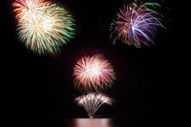 Several fireworks in a pitch black sky, launching from and reflecting off the calm ocean surface below.