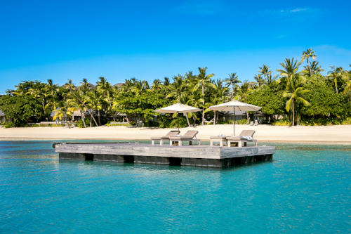 A large wooden pontoon floating on the water in front of the sunrise villas with deck chairs and umbrellas on it.