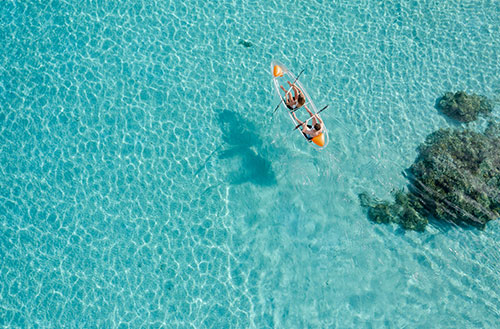 A birds eye view of a couple paddling a clear kayak through crystal clear waters at Kokomo Island Fiji.