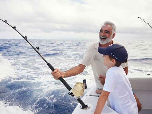 A man holding a fishing line accompanied by a young boy at the stern end of a fishing boat looking out towards the water.