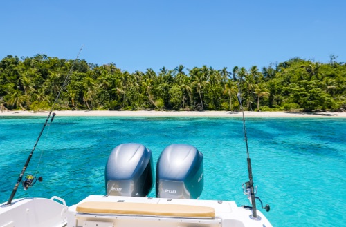 View out the back of a small boat looking at the shore of an island with two fishing rods set up to catch fish.