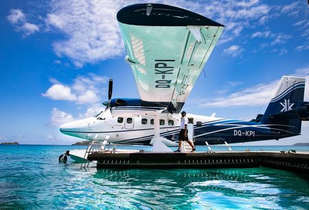 The Kokomo Private Island seaplane floating on the water while docked at a jetty, with the pilot standing next to it on the jetty on a clear, sunny day.