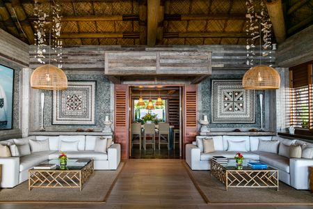 The wide anteroom of the Kokomo wine cellar, with a large dining table visible within the cellar room through the entranceway which is flanked on either side by large, modern lounge chairs and traditional Fijian artwork adorning both walls above the seating areas.