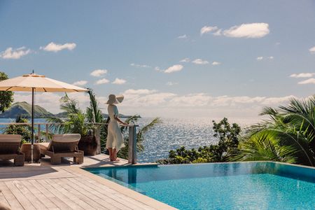 A guest standing on the wide deck of one of Kokomo’s luxury residences next to the clear infinity pool while resting against the railing, and looking out over the expansive ocean on a clear day.