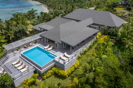 An aerial view of one of Kokomo’s hilltop residences, showcasing its large size and clear blue private infinity pool surrounded by a large tiered deck with multiple deckchairs arranged across its multiple levels. The residence is surrounded by a immaculately-maintened lawn and lush tropical vegetation.