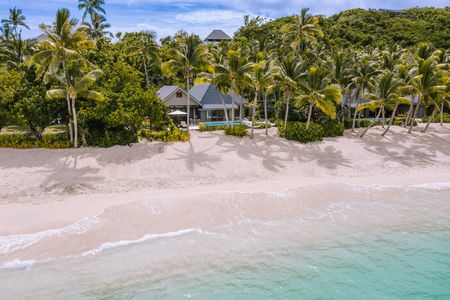 A Kokomo beachfront villa visible through palm trees and surrounded by lush vegetation, with a wide, sandy beach and clear waters directly in front of it, viewed from above the ocean water.