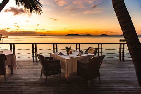 A square dining table set for dinner on the oceanfront deck of one of Kokomo Island Fiji’s restaurants. The sun is setting in the distance filling the sky with a spectrum of golden hues.