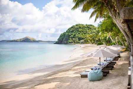 Several deckchairs shaded by large parasols lined up along a Kokomo beach, with the rest of the beach stretching away in the distance towards the end of the island running alongside the calm ocean.