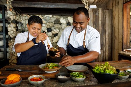 Two smiling chefs preparing a dish full of fresh produce at one of Kokomo Island Fiji’s restaurants. One chef is sprinkling seasoning on the bowl while the other drizzles honey over it.