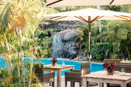 Several individual dining tables shaded by parasols across a wooden deck area next to one of Kokomo’s shared swimming pools, with a small rocky waterfall flowing into the pool seen in the distance.