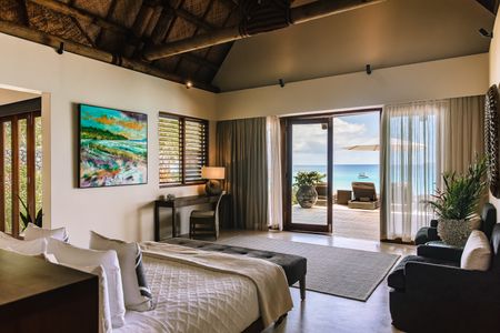 The bedroom of one of Kokomo’s beachfront villas, looking out over the large bed through the concertina doors to the wide private deck, adorned with several deck chairs facing out towards the wide open ocean where a small boat can be seen anchored nearby. A large, striking impressionist artwork is hanging inside the bedroom.