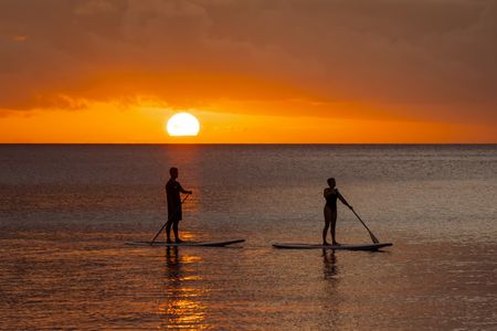 Two guests stand-up paddleboarding across the calm ocean as the glowing sun sets behind them, basking the scene in rich orange light.