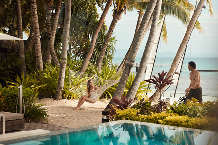 Looking out over the infinity pool at one of the Villas at Kokomo Island Fiji. With a woman laying in a hammock and a man walking out of the water with snorkelling in his hands.