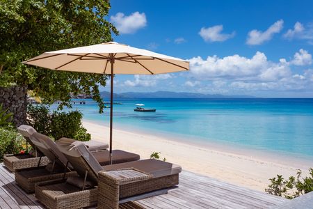 Looking out from the deck of a Kokomo beachfront villa over several private wicker deck chairs shaded by a large, white parasol with the pristine white beach and clear blue waters visible beyond them. Distant neighbouring islands can be seen on the horizon.