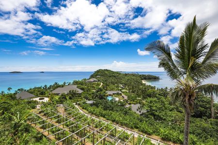 Expansive views of the entire island seen from a high vantage point, showcasing the island farm and several residences & villas, and the inland shared swimming pool visible in the centre of the island. Calm blue oceans surround the island with a few small neighbouring islands seen on the horizon to the left.