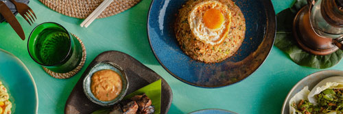 Several dishes and drinks served along a vibrant blue-green table seen from above. The dishes include skewers with satay dipping sauce, fried rice, and stuffed lettuce leaves.