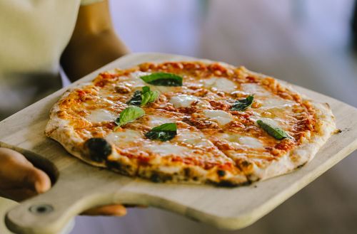 A freshly-cooked margherita pizza served on a wooden platter being carried by a waiter at Kokomo Private Island Fiji.