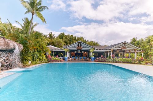 The Kokocabana Pool Club at Kokomo Private Island Fiji seen from across the wide, blue pool. A small waterfall is falling over rocks into the pool, while several deck chairs can be seen at the entrance to the Pool Club building.